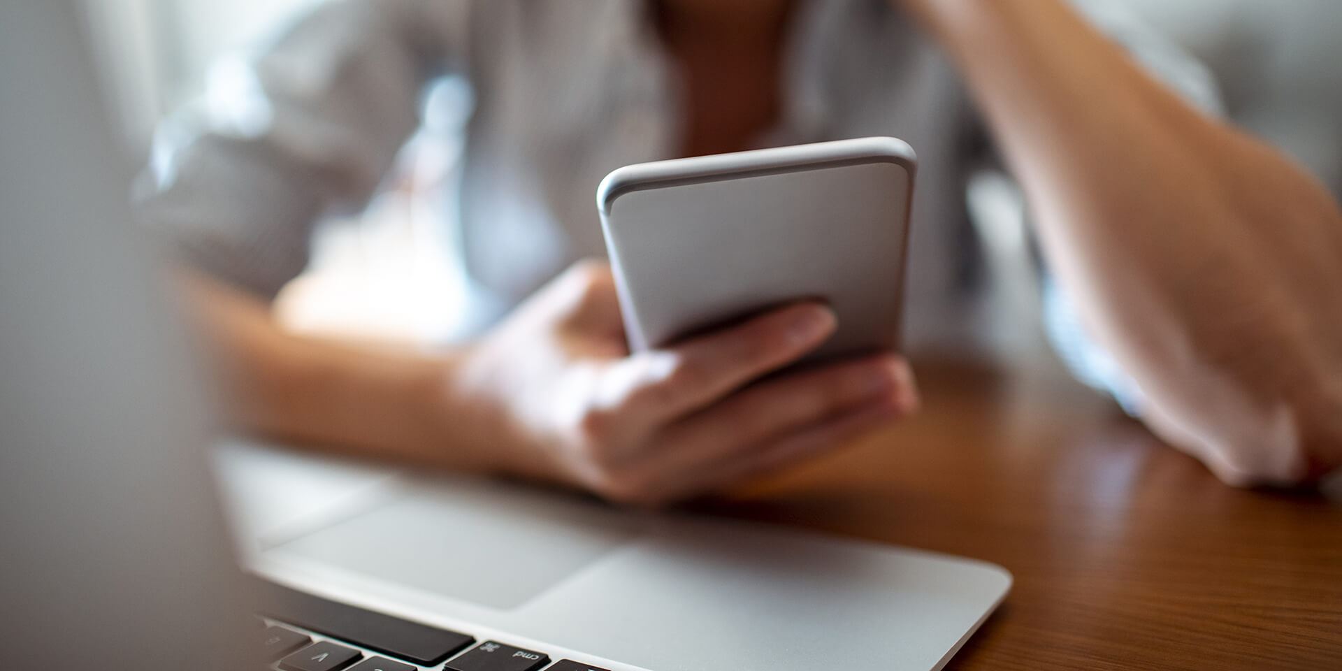 Eine Person sitzt vor einem Laptop und blickt auf ihr Smartphone